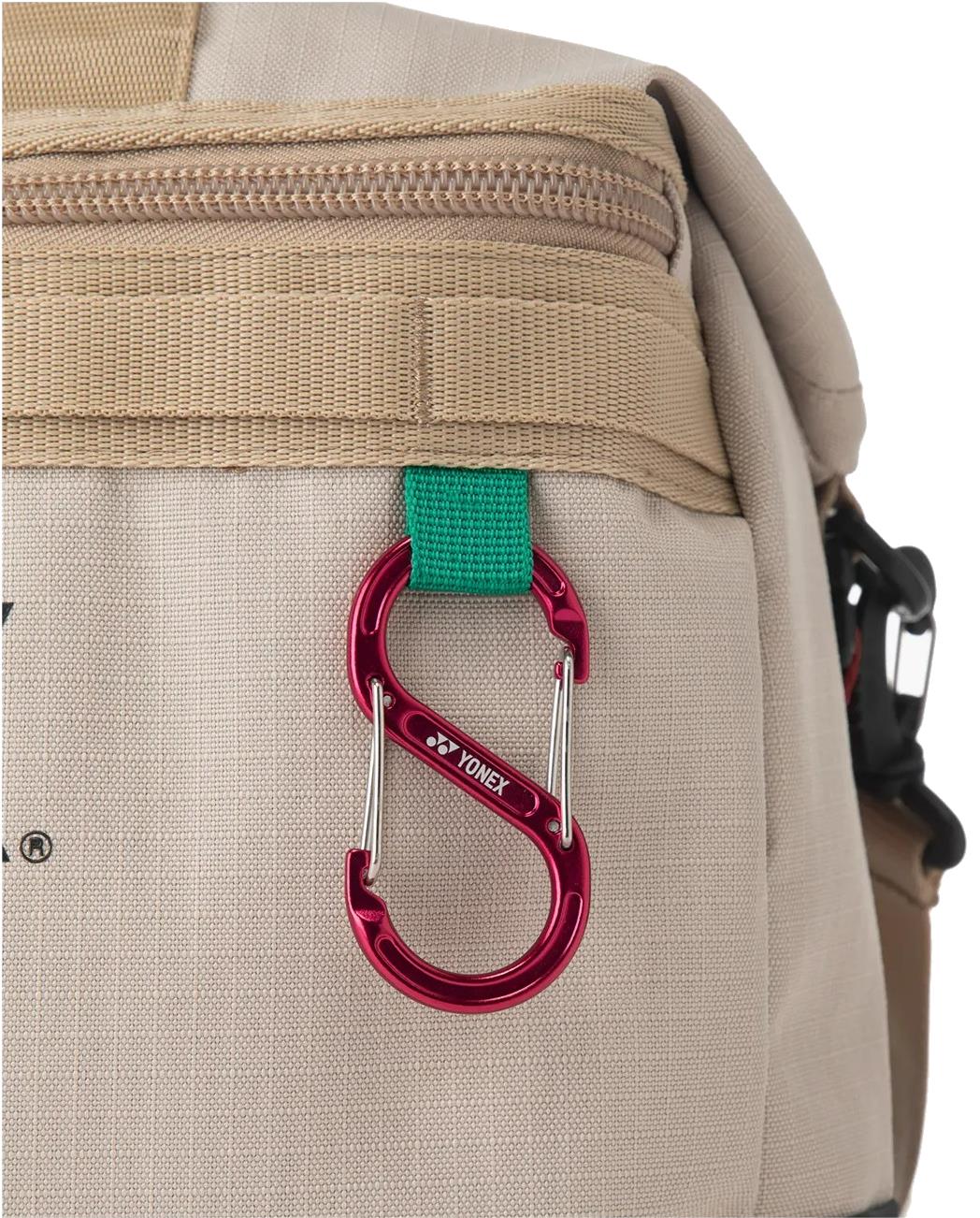 The Yonex BA72611EX Gearlogic Badminton Boston Bag in beige includes a zip pocket and features a red S-shaped Yonex carabiner on a green fabric loop, made from recycled polyester.