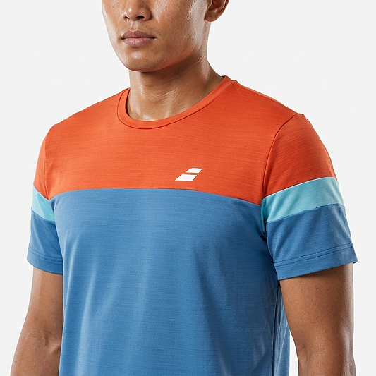 A man wearing the Babolat Crew Neck Men's Badminton T-Shirt - Seaport, featuring an athletic fit with an orange top, blue lower section, and light blue striped sleeves, stands against a plain white background.