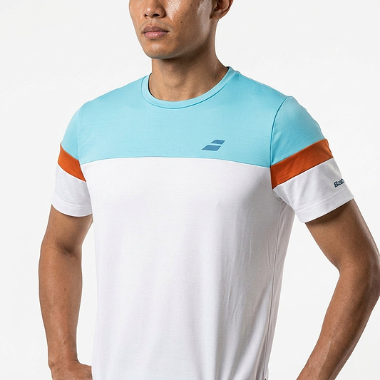 A man stands with hands on hips, wearing the Babolat Crew Neck Mens Badminton T-Shirt—white with a light blue top and orange arm stripes—against a plain background.