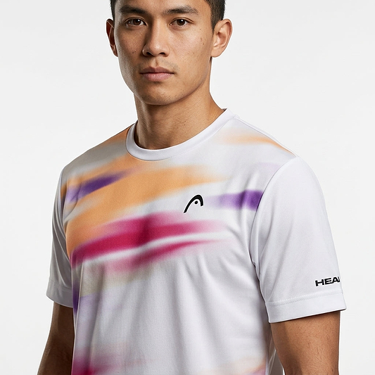 A man wears the HEAD Blur Mens Badminton T-Shirt - PUXU, a moisture-wicking tee with orange, pink, and purple blurred streaks, chest logo, and "HEAD" on the sleeve, standing against a plain white background.
