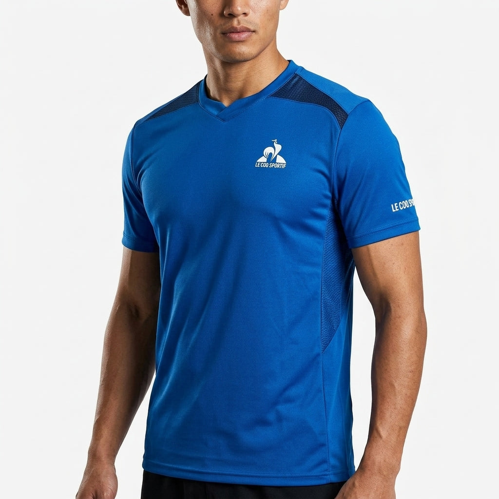 A man wears the Le Coq Sportif Pro Mens Badminton T-Shirt in Lapis Blue, featuring short sleeves, black mesh shoulder accents, and branded logos—ideal breathable sportswear from Le Coq Sportif for active days.