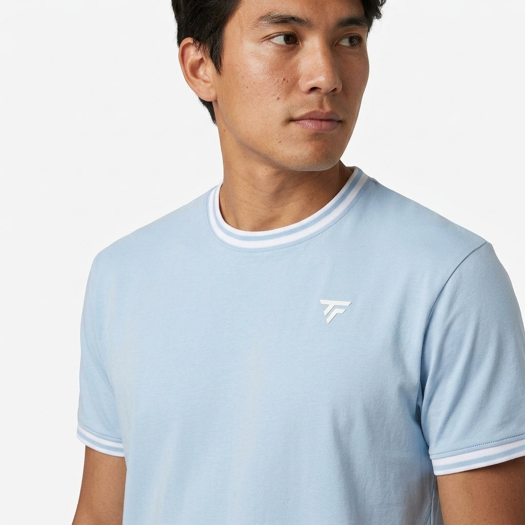 A man in a Tecnifibre Team Stretch Mens Badminton T-Shirt - Glacier Blue with white collar and sleeve trim, plus a small white chest logo, looks left. The men’s organic cotton t-shirt is shown against a plain white background.