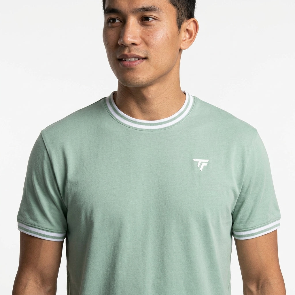 A man wears a sage green Tecnifibre Team Stretch Men's Badminton T-Shirt with white collar and sleeve trim, featuring a small white Tecnifibre logo on the left chest, standing against a plain white background.