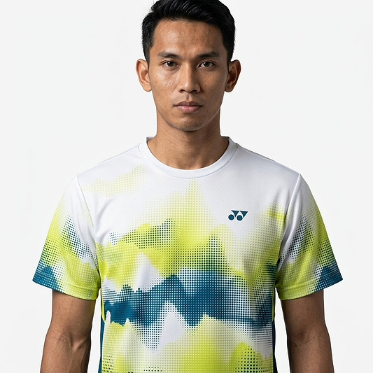 A young man with short black hair wears a Yonex YTM2025A Mens Badminton T-Shirt - White by Yonex, featuring green, blue, and yellow abstract patterns, standing against a plain white background and looking directly at the camera with a neutral expression.