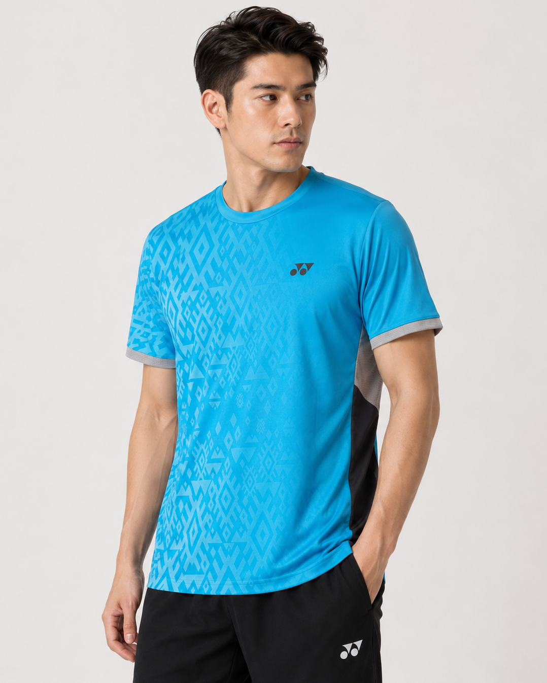 A man wears a Yonex YTM11 Men's Badminton T-Shirt in Blue Sea with a geometric pattern, paired with black shorts, his left hand in his pocket, gazing to the side against a plain light background.