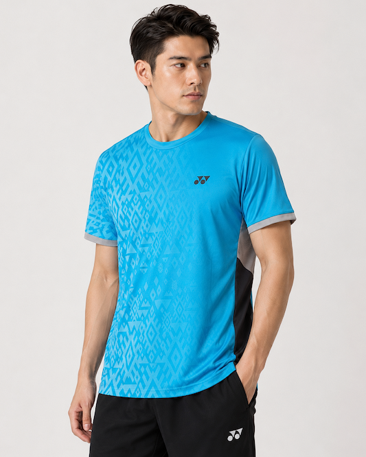 A man wears a Yonex YTM11 Men's Badminton T-Shirt in Blue Sea with a geometric pattern, paired with black shorts, his left hand in his pocket, gazing to the side against a plain light background.