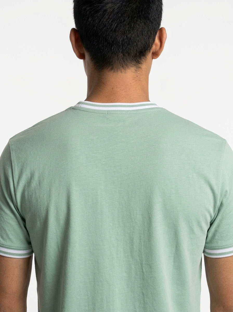 A person with short black hair is shown from behind wearing a Tecnifibre Team Stretch Men’s Badminton T-Shirt in sage green, featuring white and gray trim on the collar and sleeves, against a plain white background.