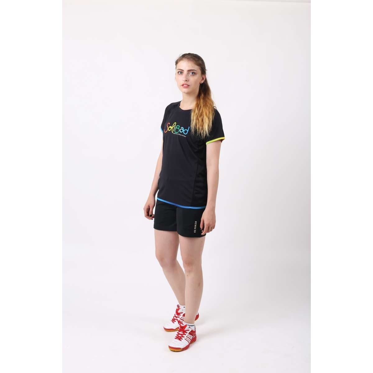 A person with long hair is in a studio setting, wearing a black FZ Forza Bulgur Solibad Badminton T-shirt, black athletic shorts, and red and white athletic shoes. The plain white backdrop highlights their connection to the badminton community.