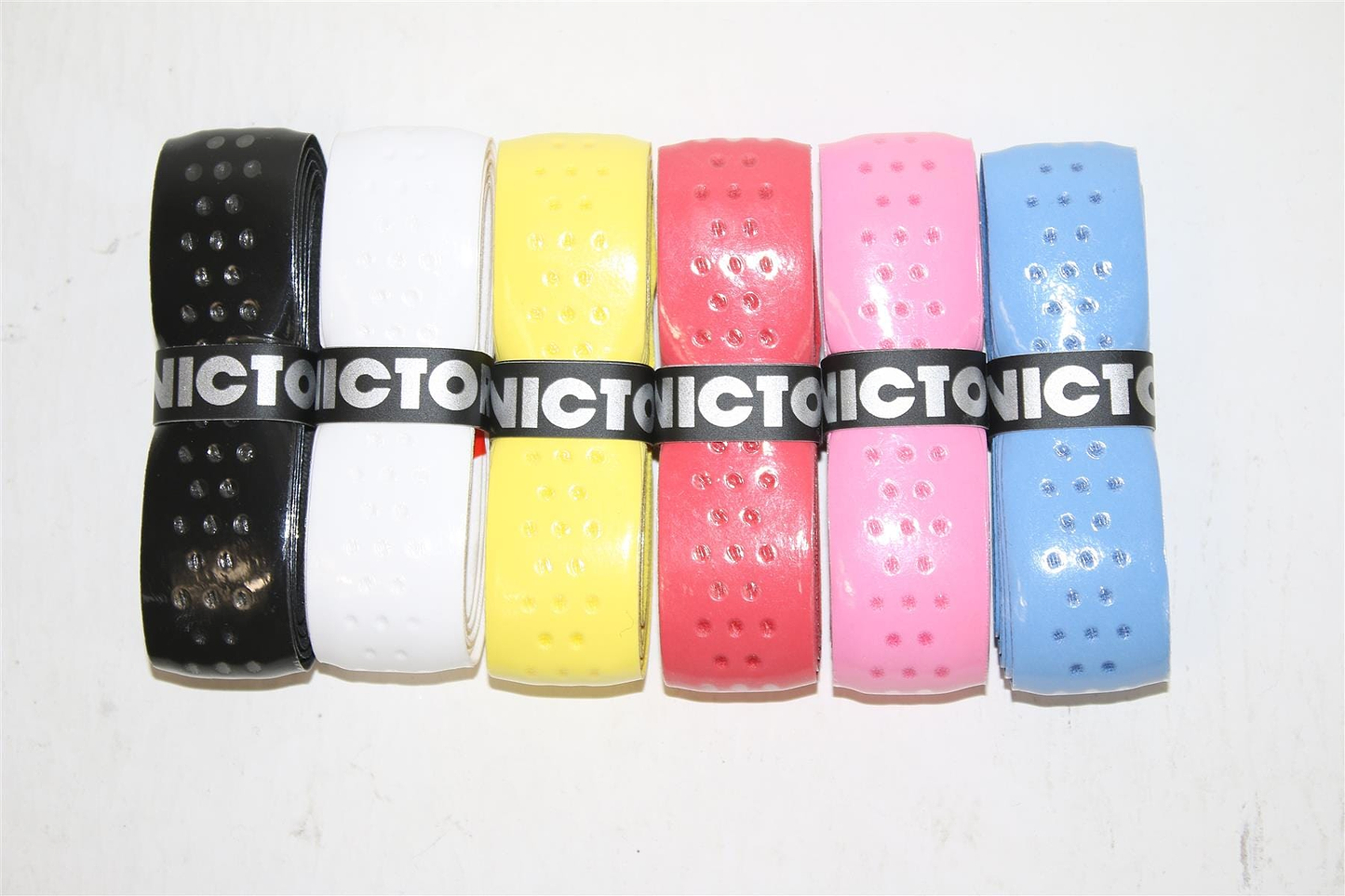 A lineup of six vividly colored Victor Soft Grip Replacement Badminton Grips, akin to those used for bicycle handlebars, features perforated edges and is held together by black bands with white text. The array of colors—black, white, yellow, red, pink, and blue—is set strikingly against a crisp white backdrop.