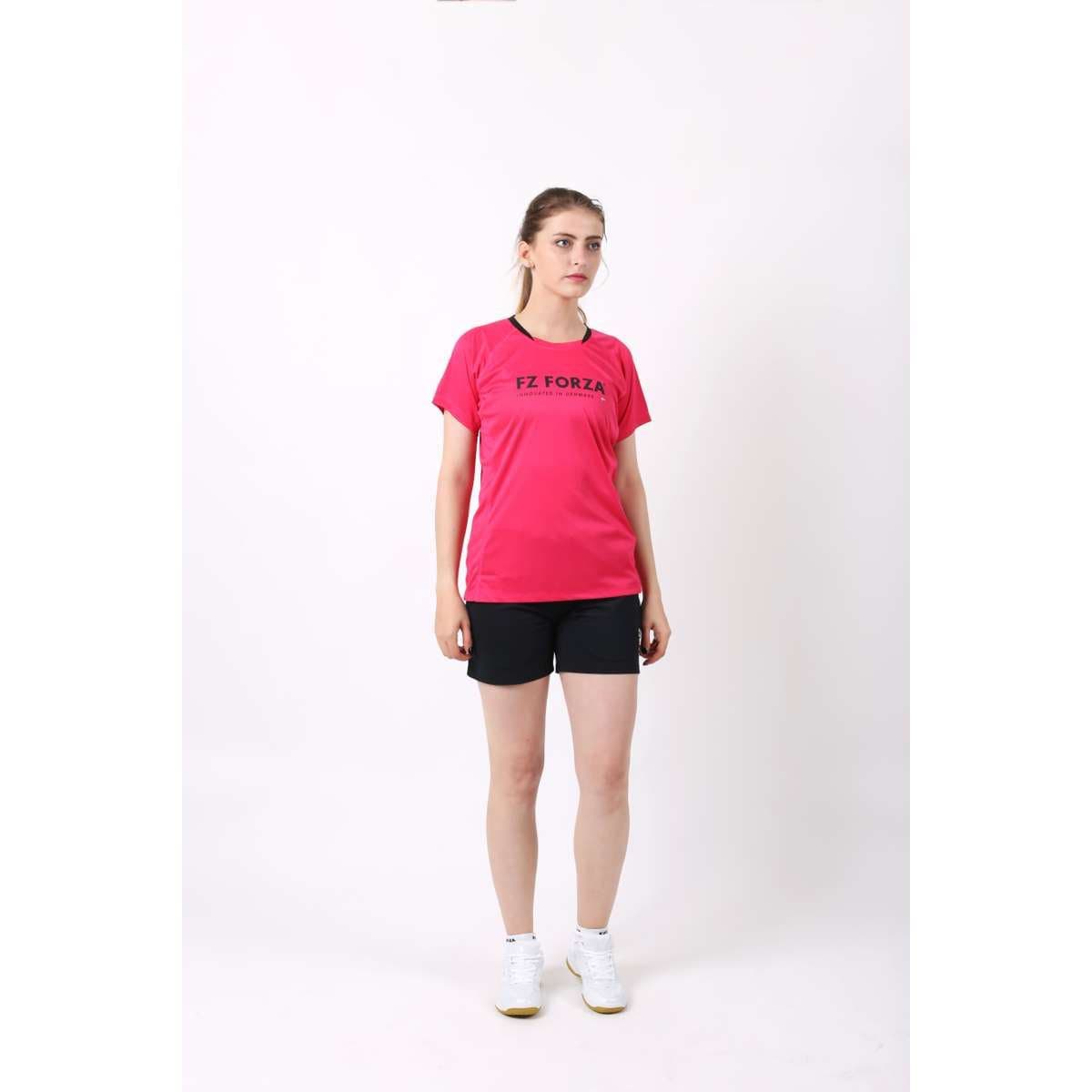 A person stands against a white background, wearing a bright pink FZ Forza Blingley Sparkling Cosmo Pink Badminton T-Shirt, made of quick-drying material. They pair it with black shorts and white sneakers. Their long hair is tied back as they look slightly to the side, ready for a game.