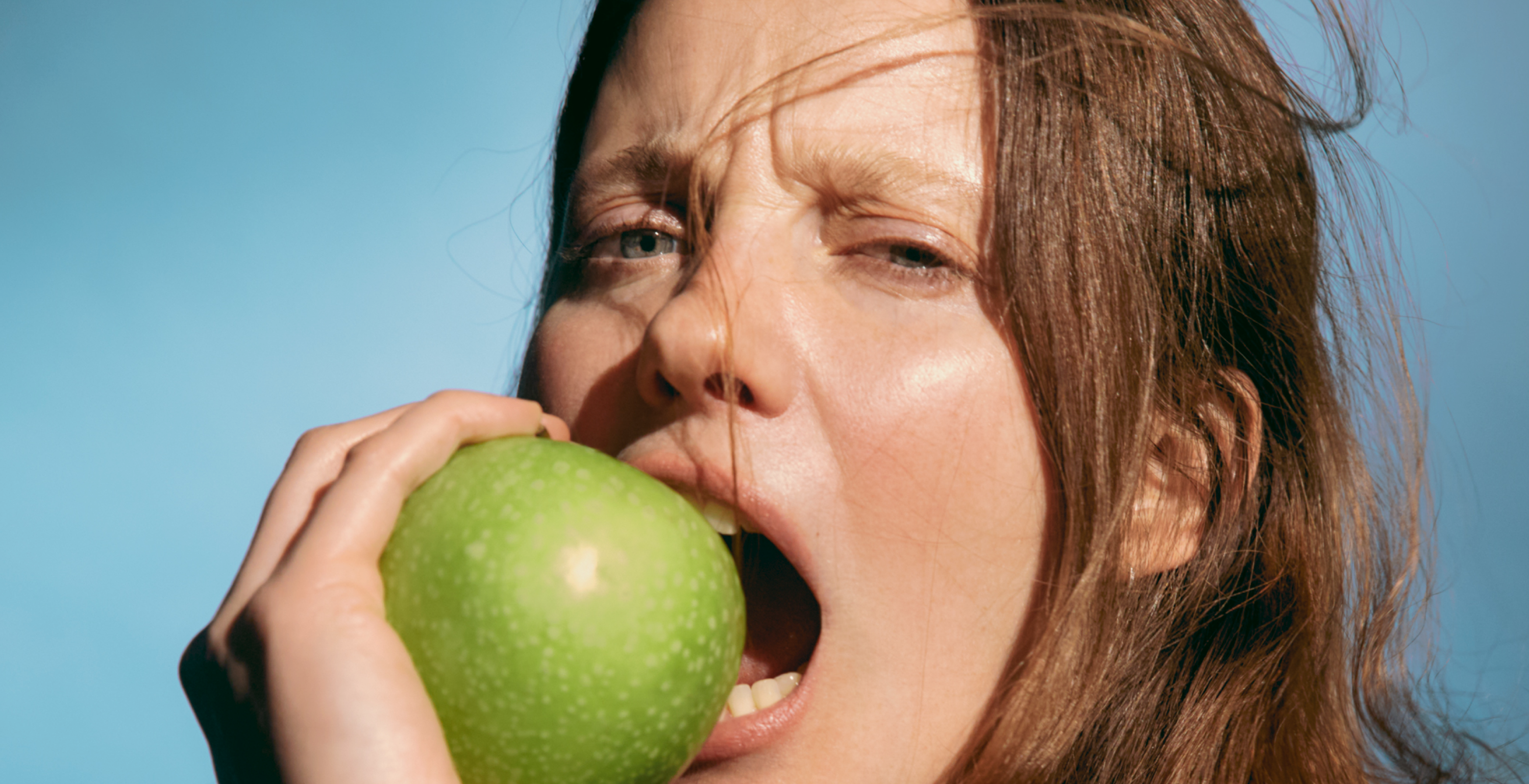 Close-up of a woman biting a green apple against a blue sky, symbolizing freshness and natural vitality