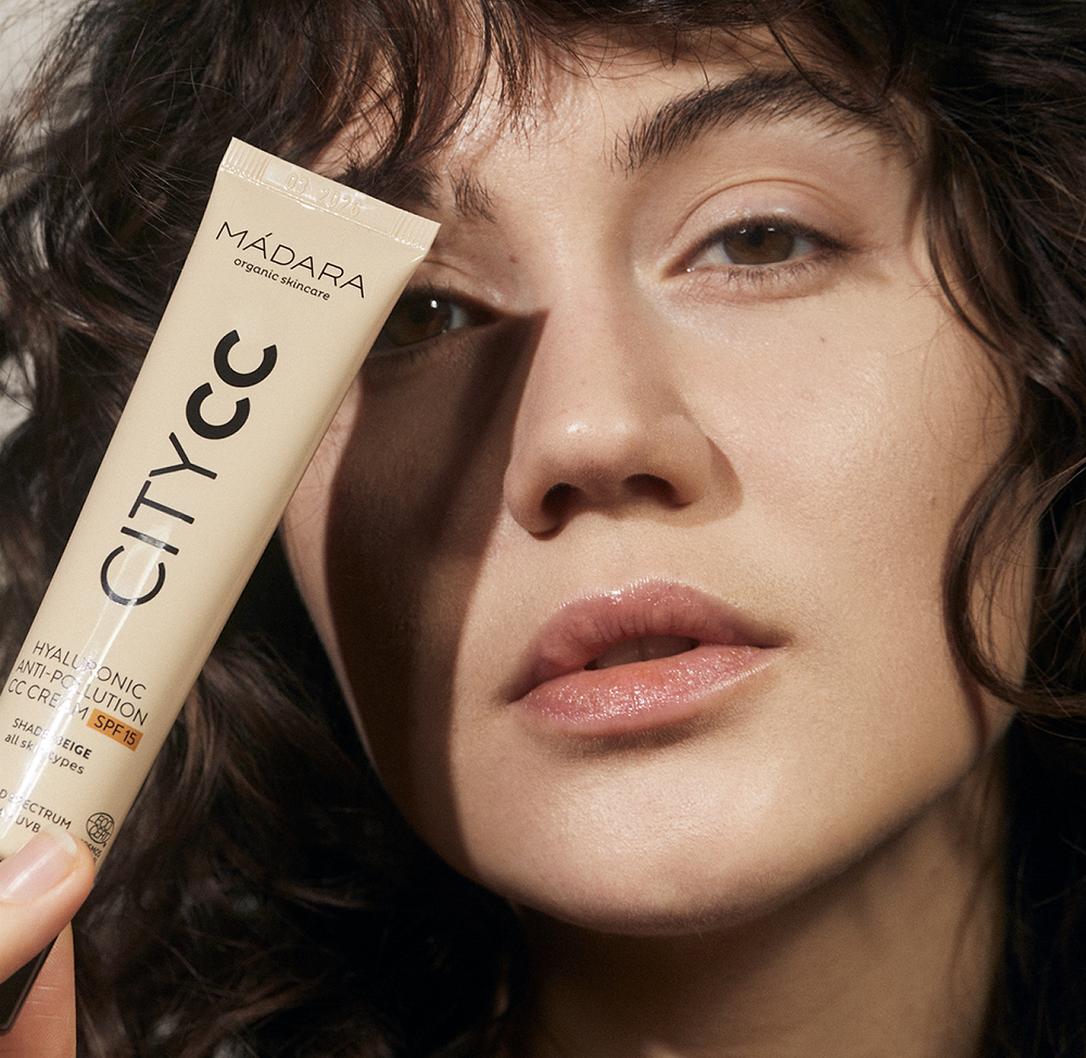 Model holding MÁDARA CITY CC Hyaluronic Anti-Pollution CC Cream SPF15 tube beside her face, showing natural glowing skin and lightweight complexion coverage