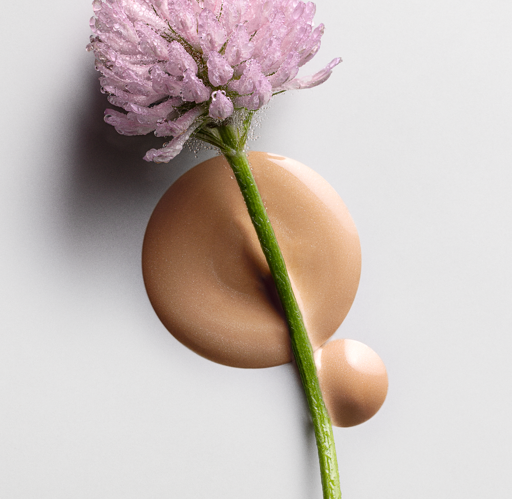 Close-up of a dew-covered pink clover flower placed over smooth drops of beige tinted serum on a light background, representing the texture of MÁDARA Cosmetics H2O Hyaluron Tinted Serum in Sun Flower shade