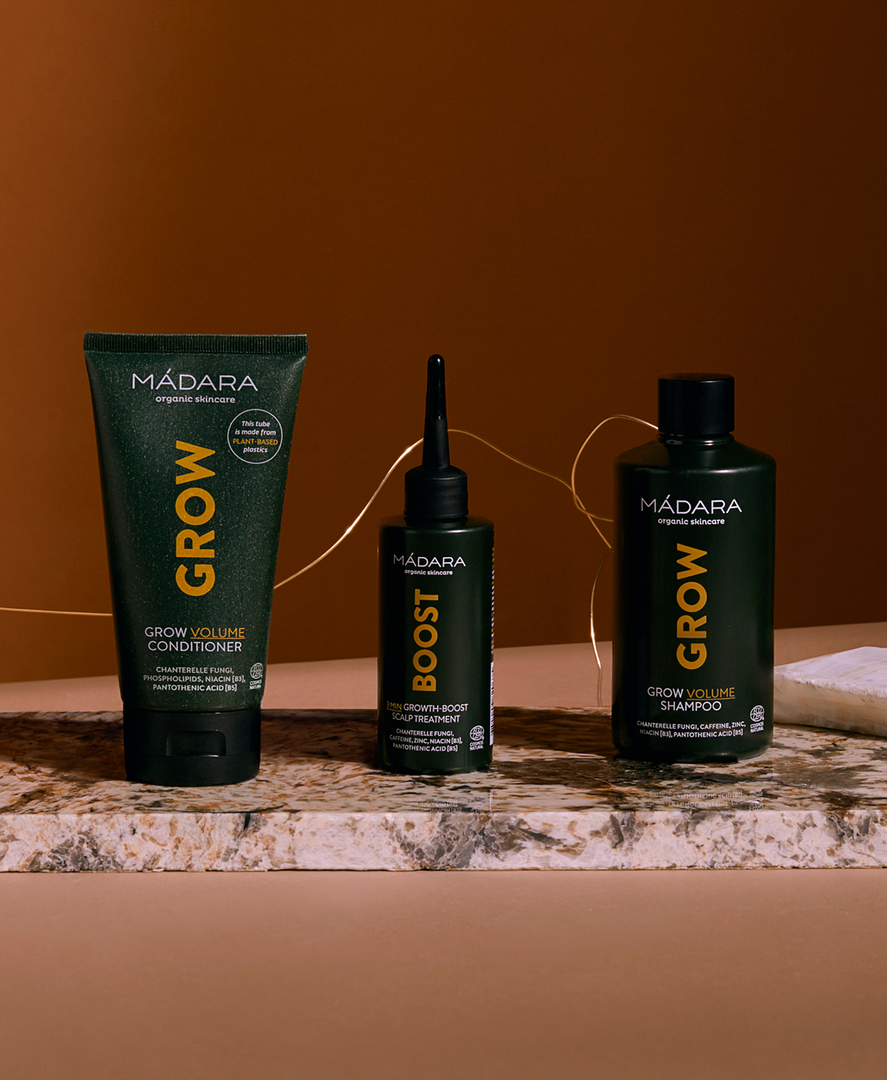 MÁDARA Cosmetics Grow haircare line displayed on a marble surface against a warm brown background, featuring Grow Volume Conditioner, Boost scalp treatment and Grow Volume Shampoo