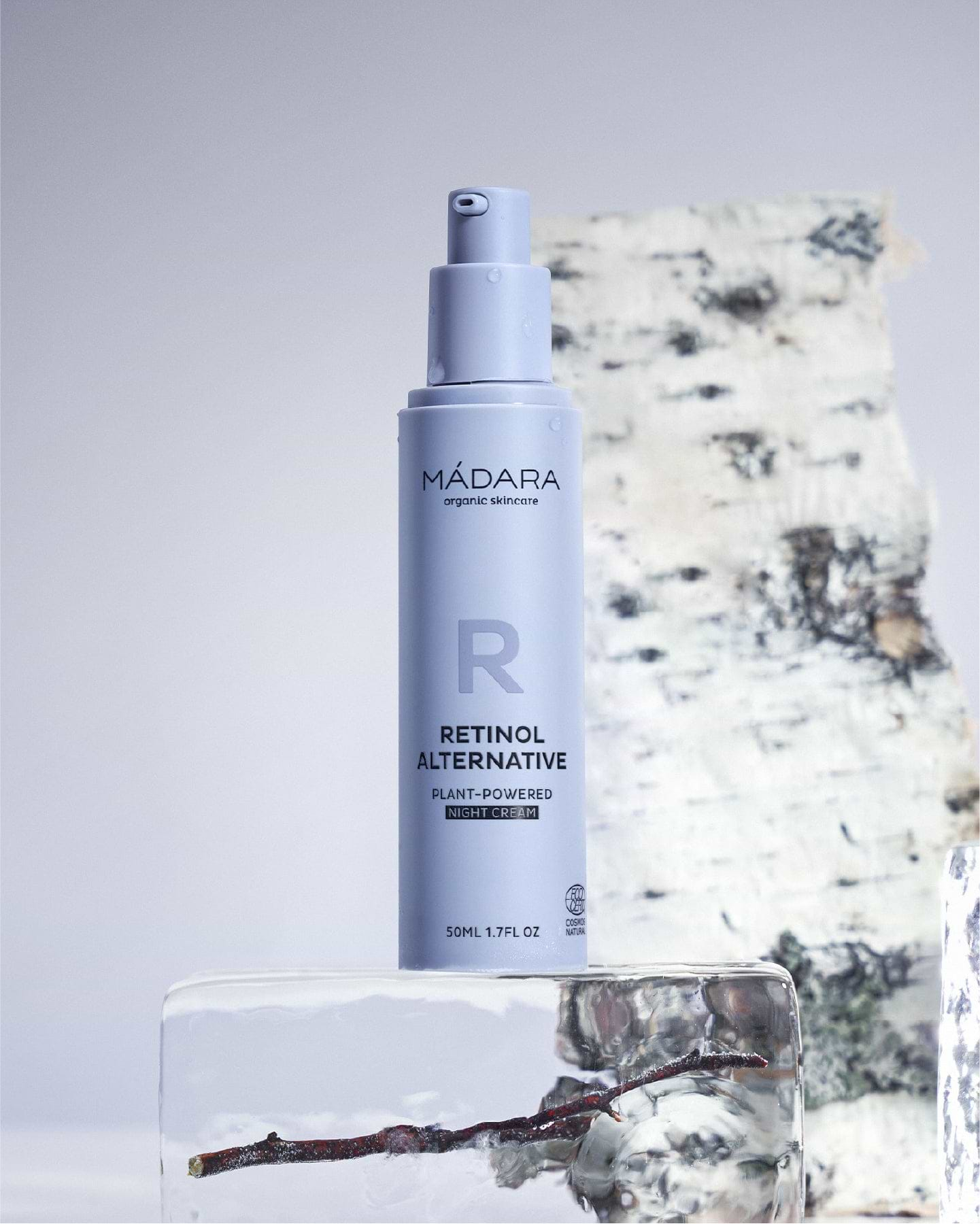 A 50 ml pump bottle of Madara Organic Skincare Retinol Alternative Plant-Powered Night Cream without the plastic cap, placed on a block of ice and a birch tree log in the background