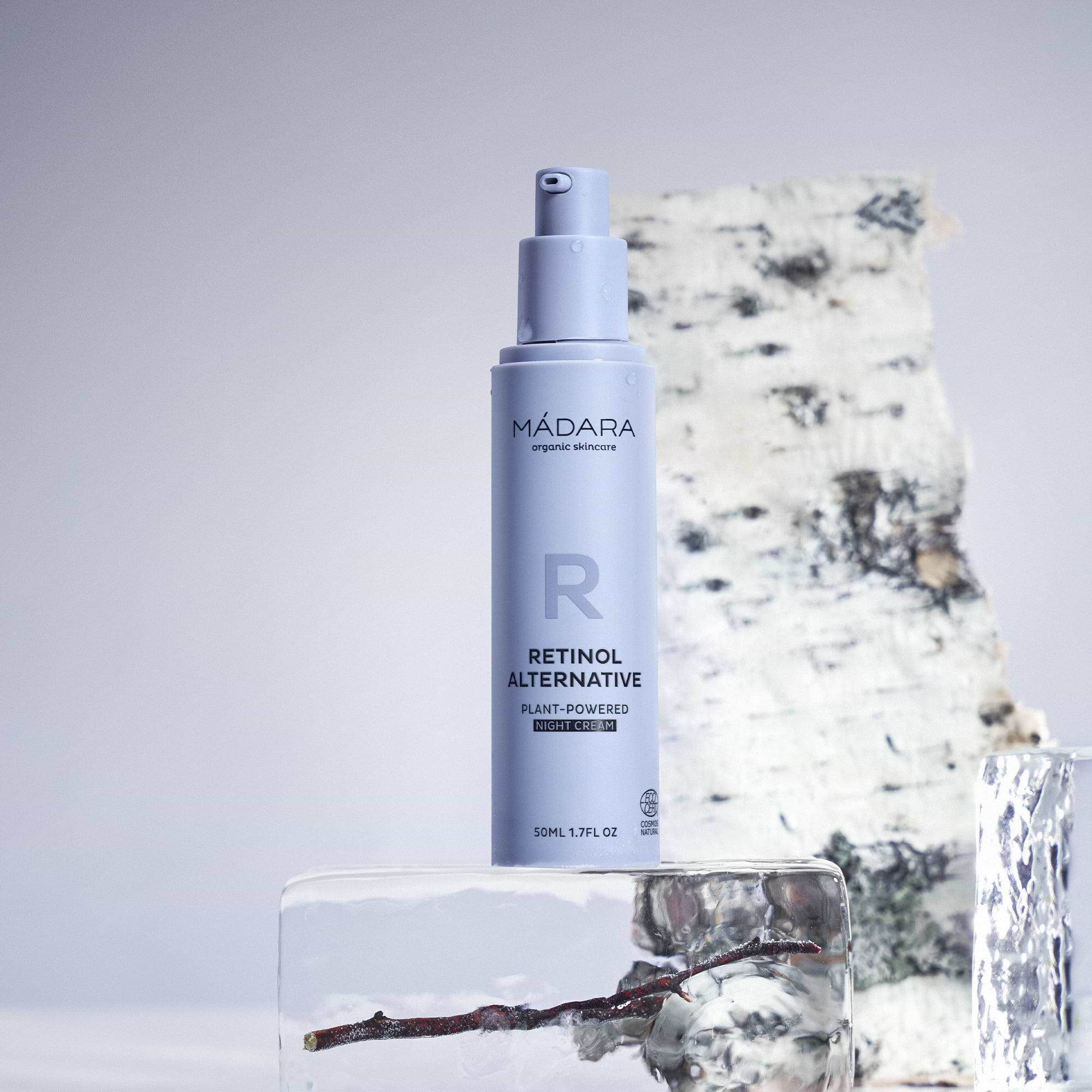 A 50 ml pump bottle of Madara Organic Skincare Retinol Alternative Plant-Powered Night Cream without the plastic cap, placed on a block of ice and a birch tree log in the background