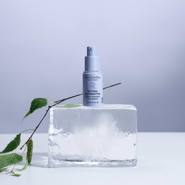 A 15 ml pump bottle of Madara Organic Skincare retinol alternative plant-powered eye serum with no cap, placed on a block of ice next to a small branch of birch tree