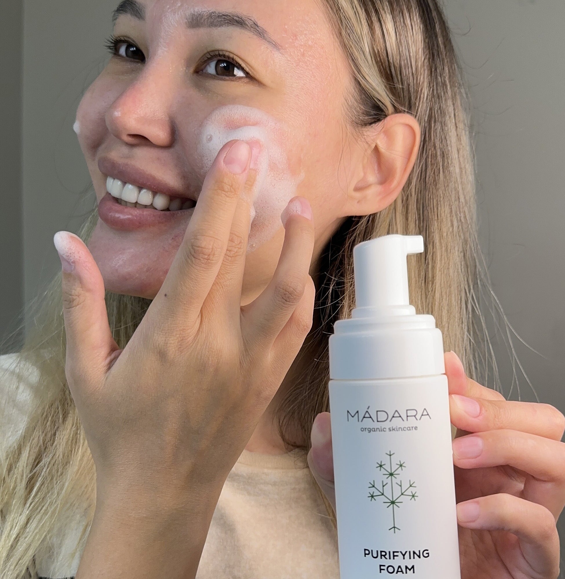 Woman holding a bottle of Mádara Cosmetics Purifying Foam for face cleansing while applying the foam to her face.