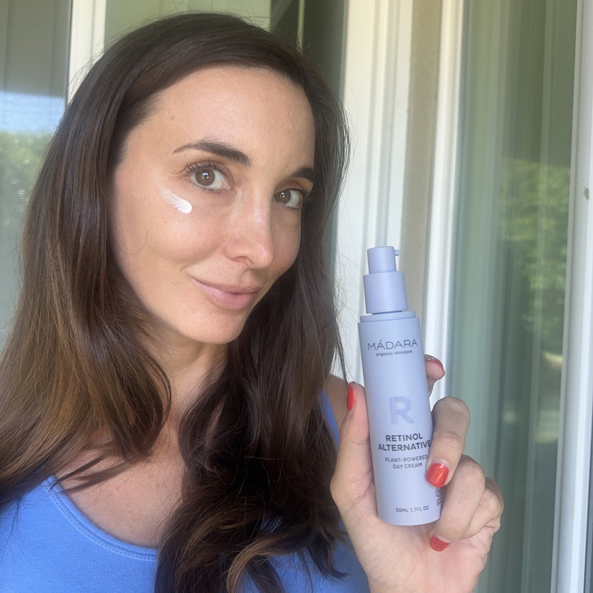 Woman holding a bottle of Mádara Retinol Alternative Day Cream with cream aplyed to her face, promoting plant-powered skincare for anti-aging benefits.