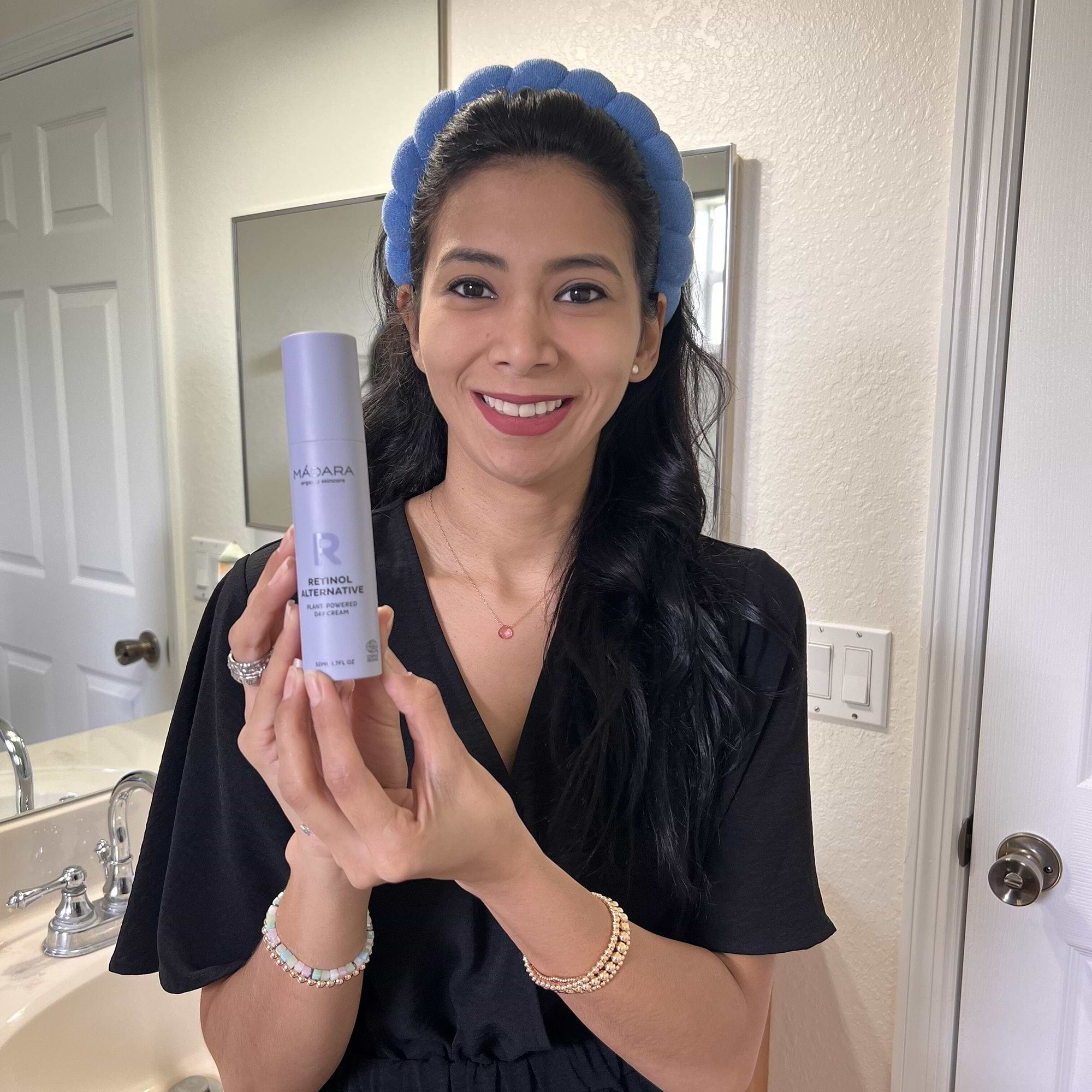 Woman smiling and holding a bottle of Mádara Cosmetics Retinol Alternative Cream in a bathroom, showcasing skincare product for a natural anti-aging routine