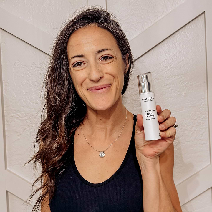 Smiling woman holding a bottle of Mádara Time Miracle Total Renewal Night Cream, showcasing anti-aging skincare for overnight rejuvenation.