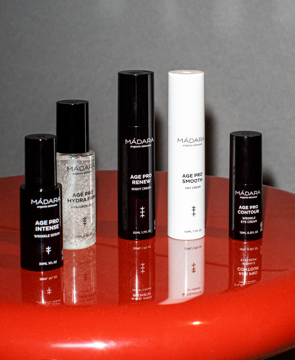 Five MÁDARA Age Pro skincare products—serum, hydra firm jelly, night cream, day cream, and eye cream—arranged on a reflective red surface against a neutral background
