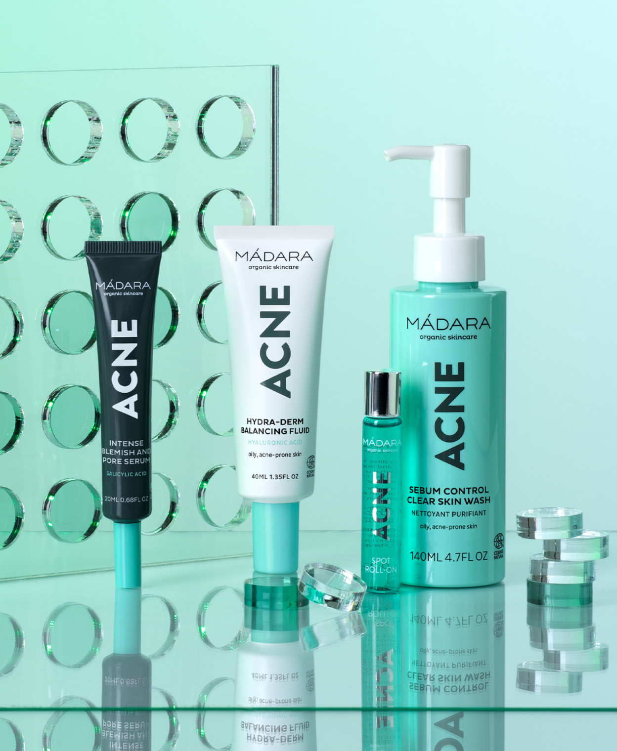 MÁDARA ACNE skincare range including blemish serum, balancing fluid, spot roll-on, and clear skin wash, displayed on a reflective surface with a mint-green background and glass elements