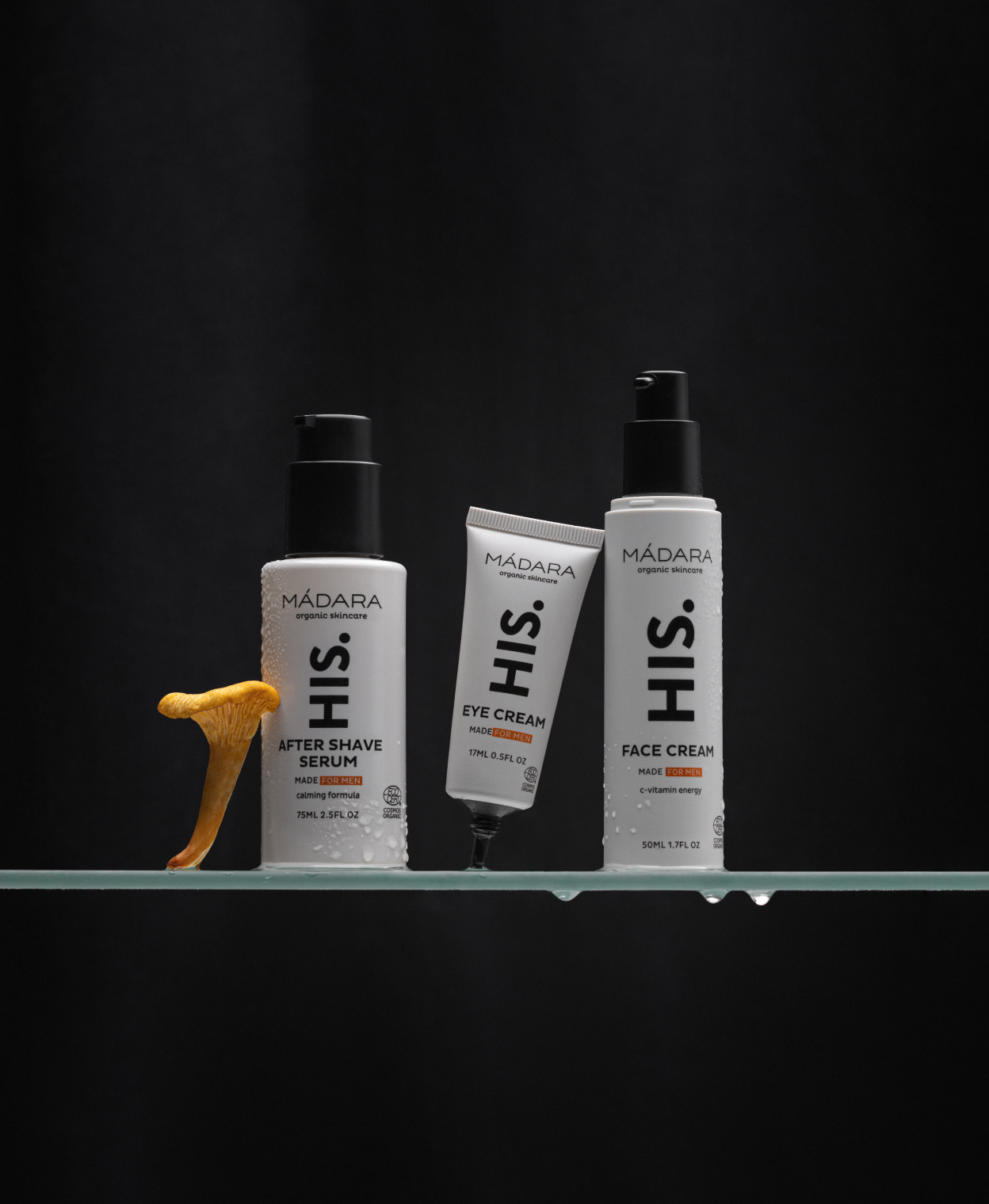Three MÁDARA HIS skincare products—after shave serum, eye cream, and face cream—displayed on a glass shelf with water droplets and a chanterelle mushroom against a dark background