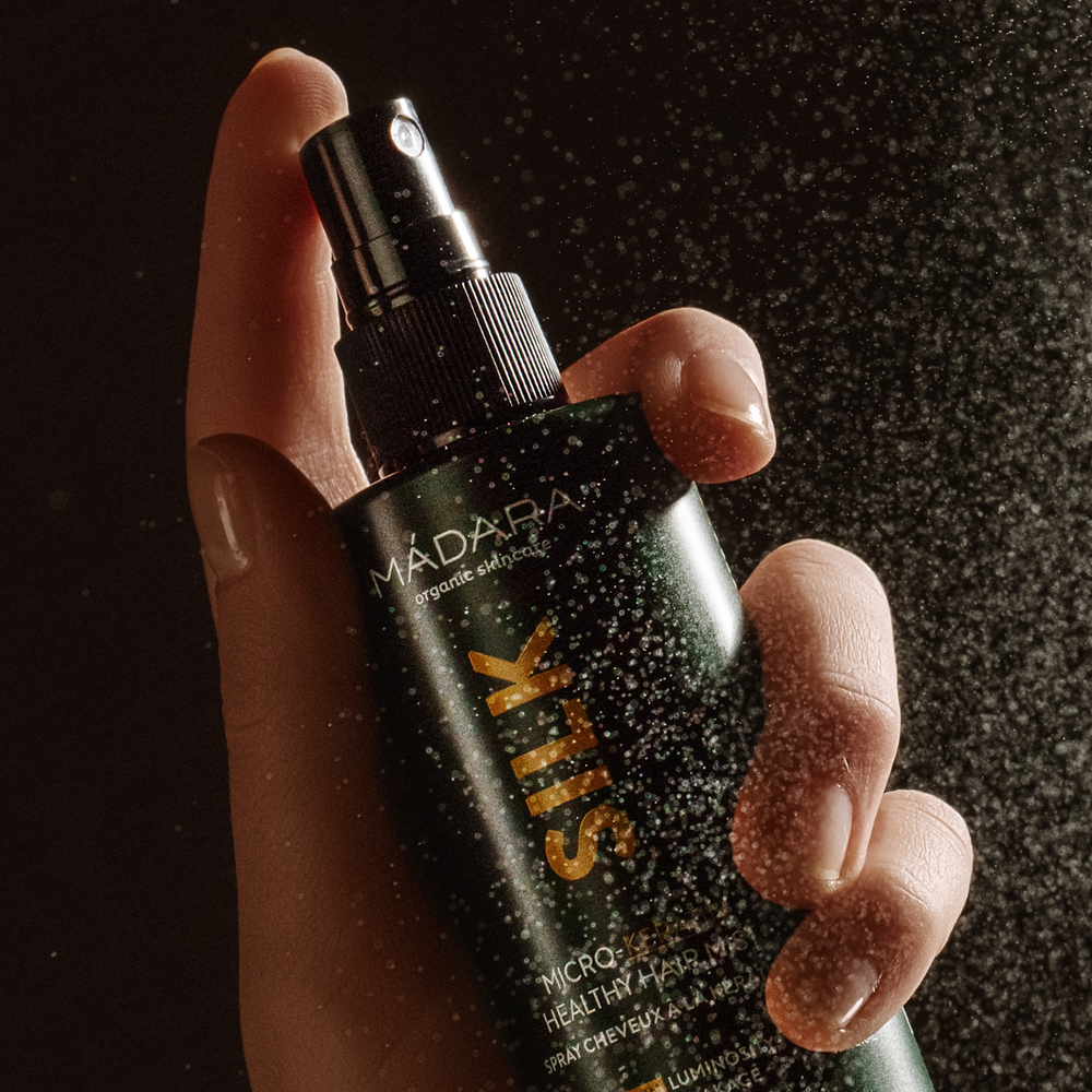 Close-up of a hand spraying MÁDARA Silk Micro-Keratin Healthy Hair Mist, with fine mist particles visible in warm lighting