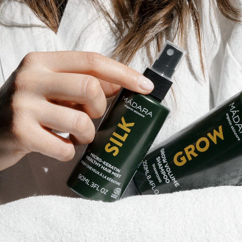 Hand holding MÁDARA Silk Micro-Keratin Healthy Hair Mist alongside Grow Volume Shampoo, both in green packaging