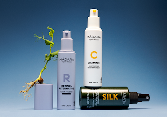 MÁDARA skincare products displayed on a blue background with a retinol alternative serum, Vitamin C cream, a silk hair product, and a small plant growing from a container