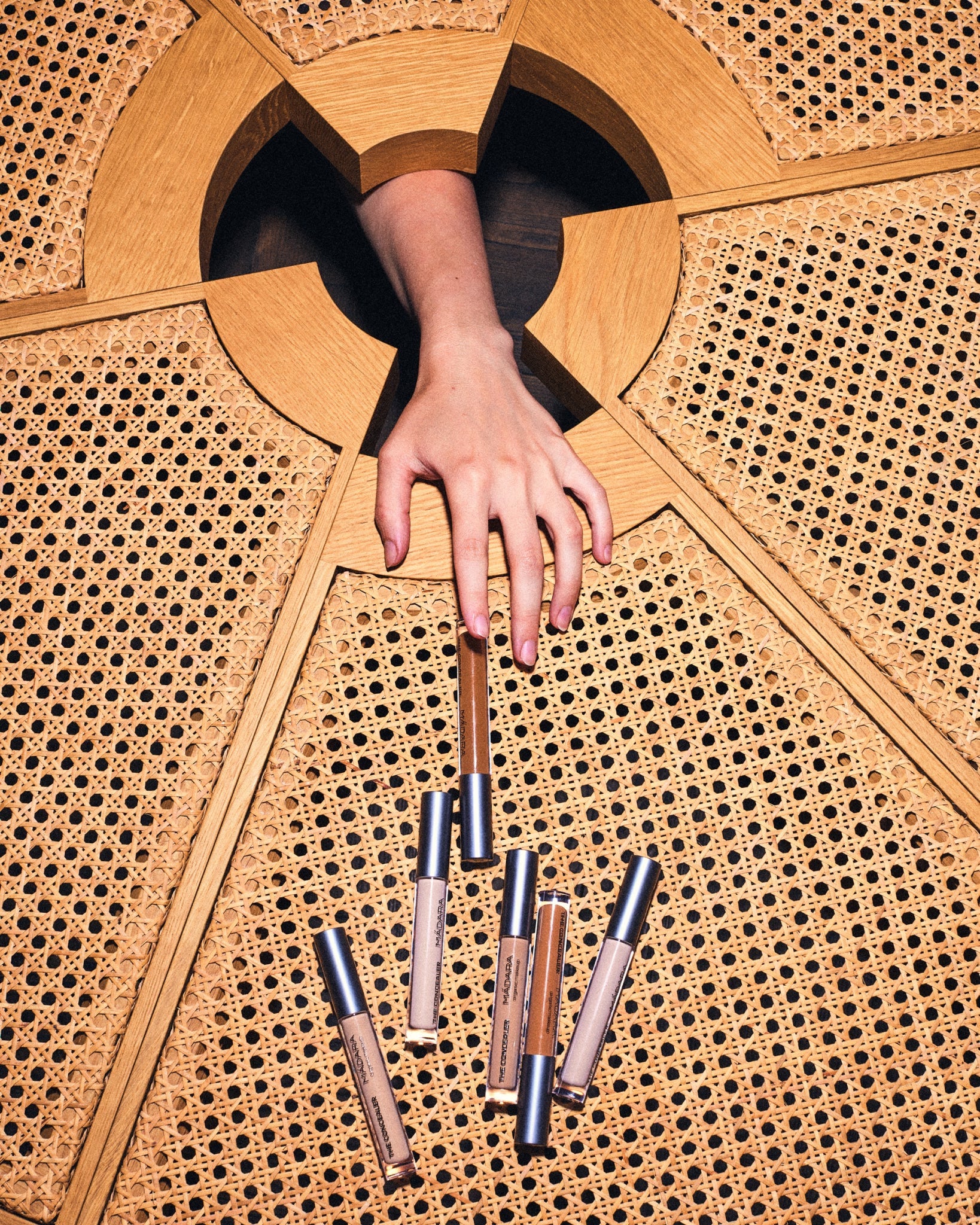 A hand reaching through a wooden design to get MÁDARA concealer on a woven rattan surface.