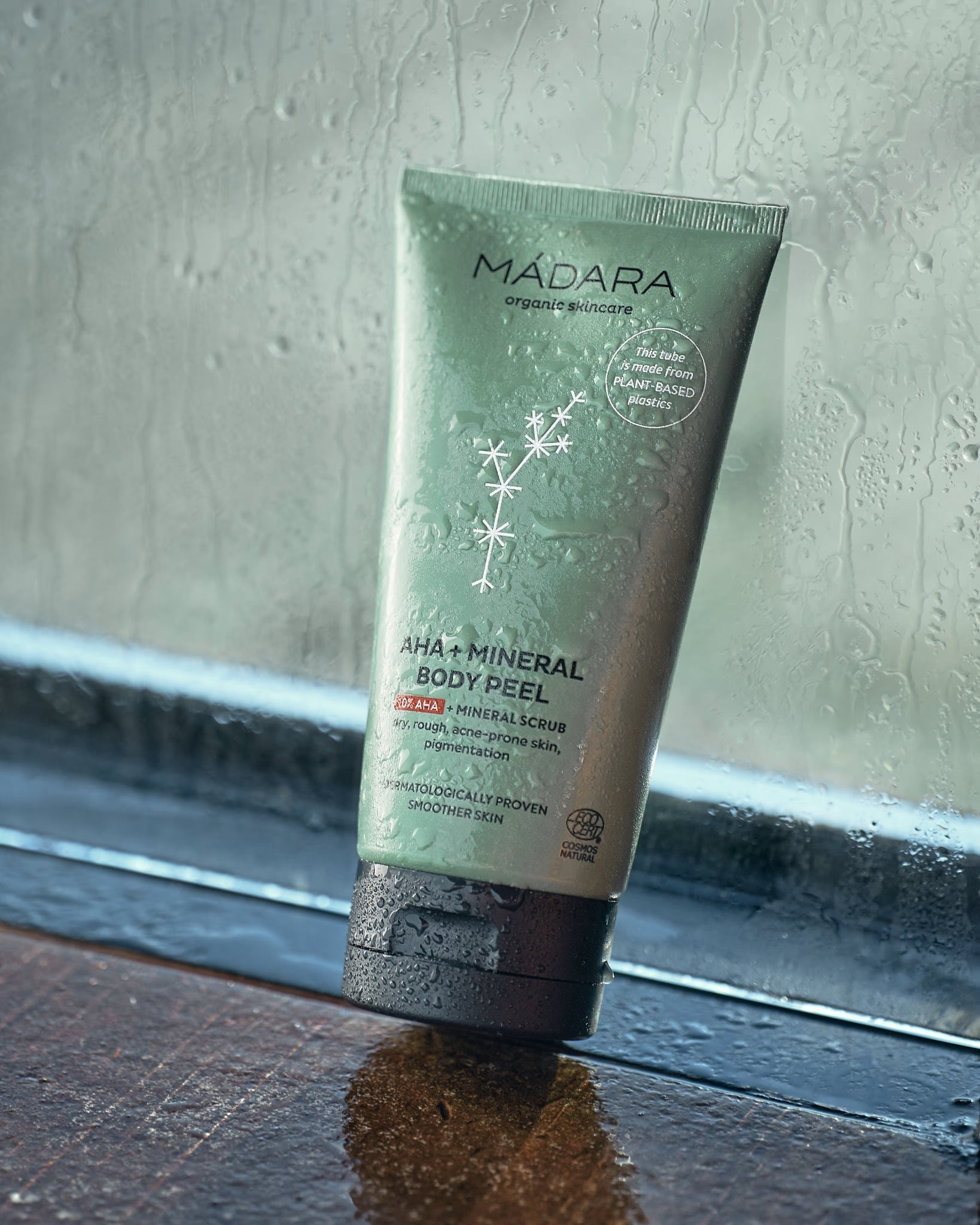 Madara Organic Skincare AHA + Mineral Body Peel resting on a dark ledge by a rain-streaked window.