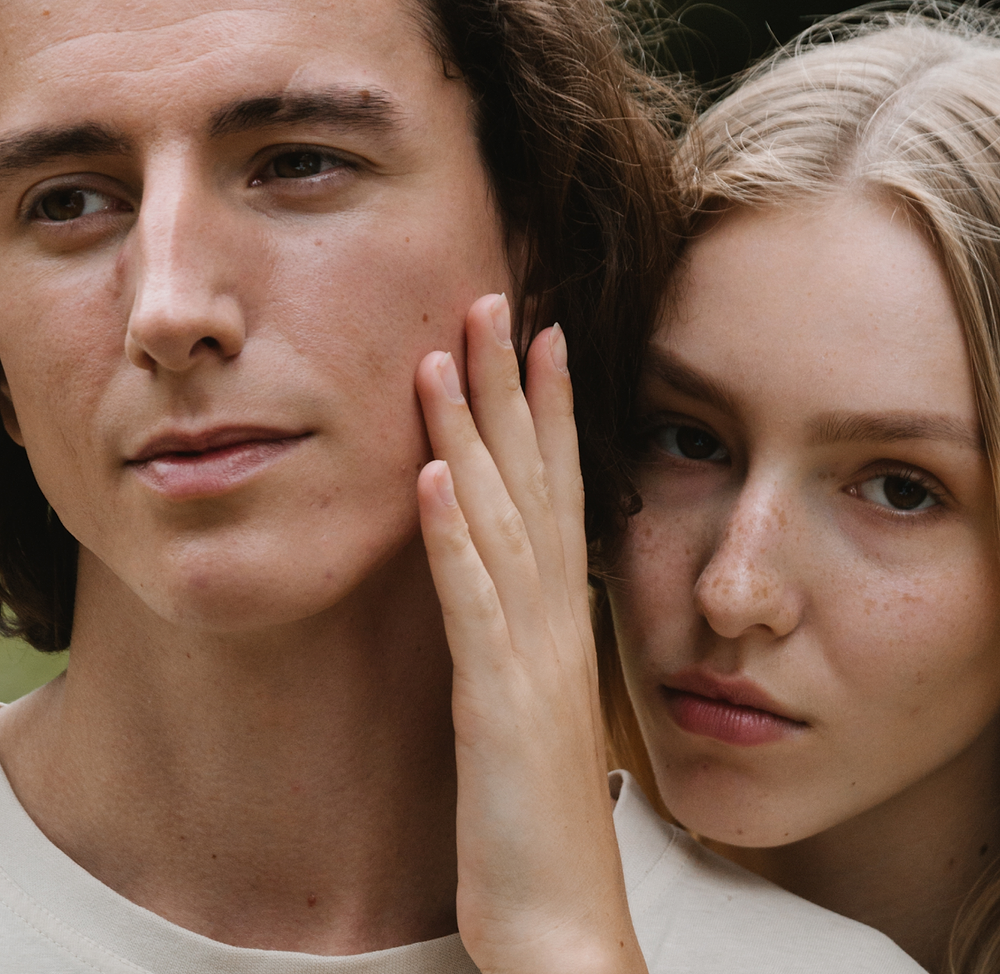 close-up of two young adults with natural, blemish-prone skin, one gently touching the other’s cheek, showcasing clear skin texture — while using the madara acne line
