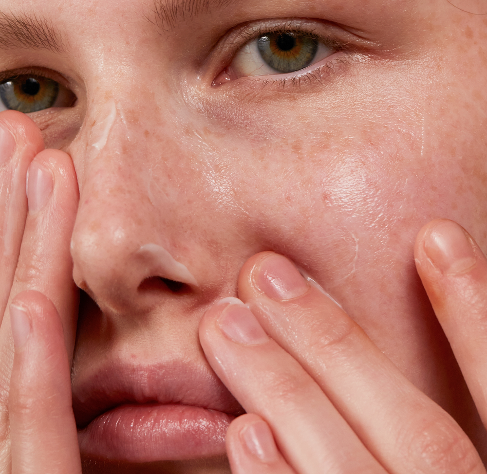 Close-up of a woman applying madara derma collagen skincare cream to hydrated, glowing facial skin with visible freckles and natural texture