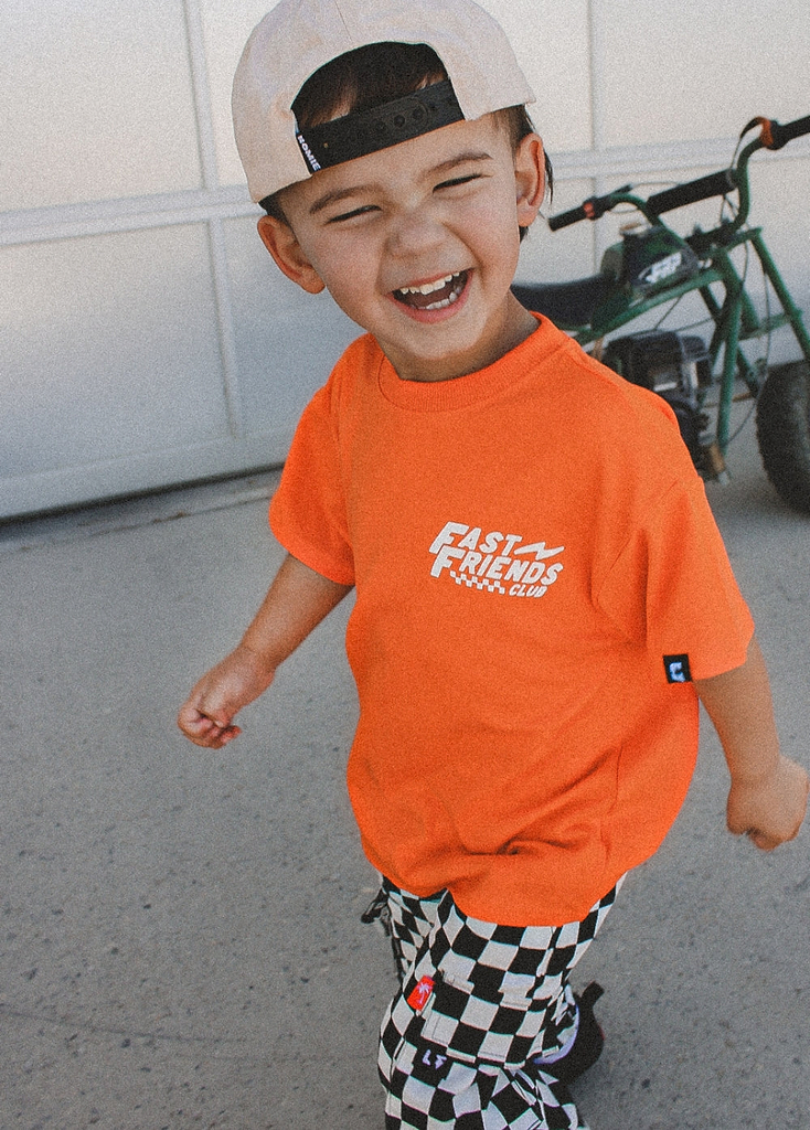 Child wearing an orange t-shirt with text, black and white checkered pants, and a white cap outdoors.