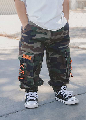 Boy wearing Little Trouble camouflage cargo pants with orange accents and black sneakers on a concrete surface.