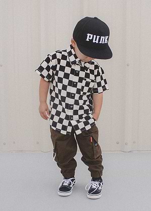 Child wearing a Little Trouble outfit. Checkered button down short sleeve shirt, brown pants, and a black hat against a plain background.