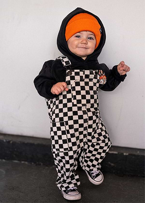 Child wearing a black hoodie, orange beanie, and Little Trouble Checkered Overalls against a white wall.