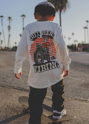 Person wearing a Out Here Vibin Long Sleeve on the back, standing on a beach with palm trees in the background.