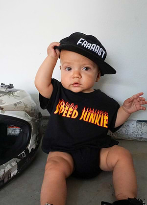 Baby wearing a Little Trouble black Snapback hat and onesie Speed Junkie Baby Romper, sitting on a concrete floor.