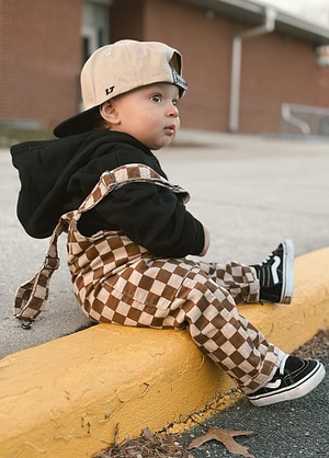 Child wearing a black hoodie, beige teddy checker overalls, and black sneakers sitting on a yellow curb.