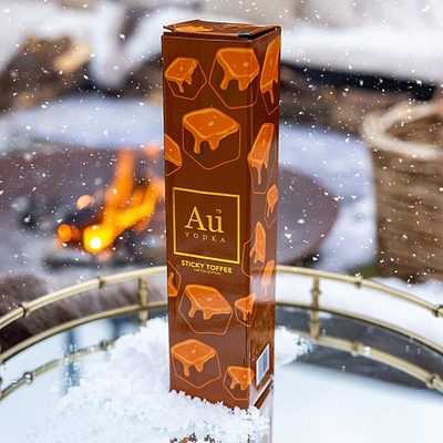Au Vodka Sticky Toffee Collection Box packaging in front of a snowy outdoor scene with a fire.