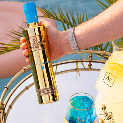 Person Holding a Bottle of Au Vodka Blue Raspberry 375ml, next to a Au Diamond Cut Glass full of blue liquid. On a reflective surface, next to an outdoor pool.