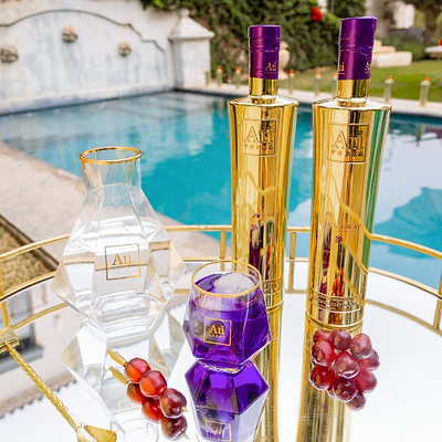 Two Bottles of Au Vodka Black Grape, next to a clear decanter & diamond cut glass with purple liquid. Set on a reflective surface, next to an outdoor pool backdrop. 