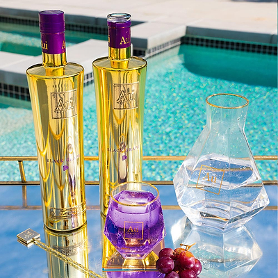 Two bottles of Au Vodka Black Grape alongside a clear decanter, diamond cut glass with purple liquid and purple grapes, set against a poolside backdrop.