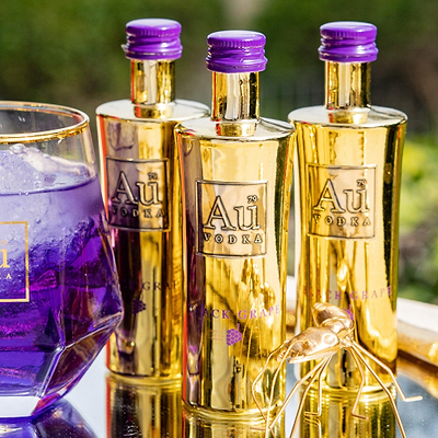 Three miniature bottles of Au Vodka Black Grape with gold-colored labels and purple caps, displayed alongside a glass of purple liquid.