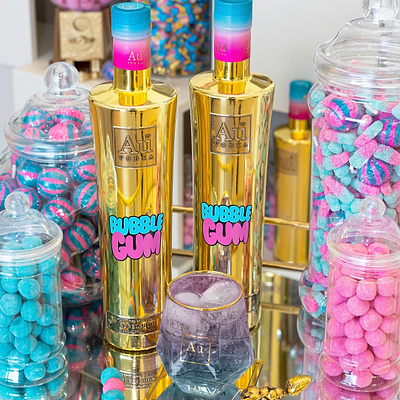 Two Bottles of Au Vodka Bubblegum close up, next to a Diamond Cut Glass, filled with ice and bubblegum liquid. surrounded by jars of candy. 