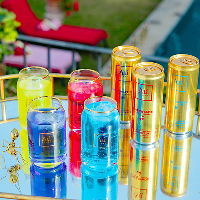 A set of colorful cocktail-style drinks in clear glasses and gold cans labeled 'Au' and 'Cherryade', arranged on a mirrored tray outdoors near a pool and lounge chair.