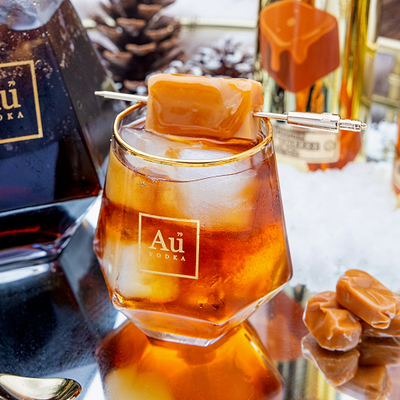 Au Vodka Sticky Toffee with ice cubes and a cocktail stick in a Diamond Cut glass labeled 'Au Vodka', surrounded by bottles and caramel candies.